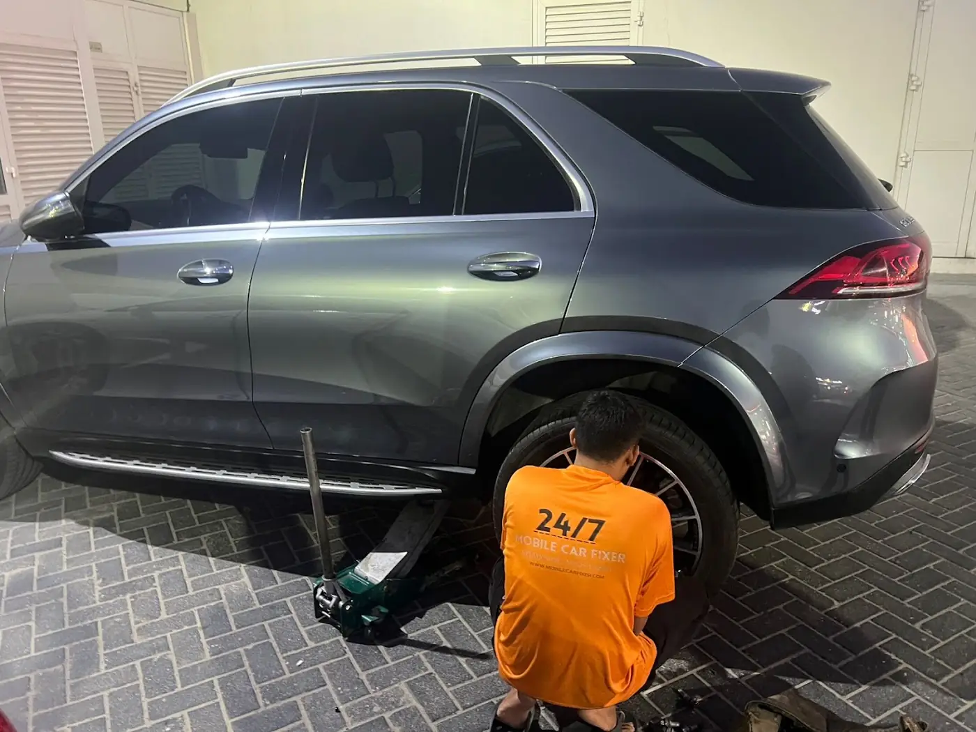 A Mobile Tire Fixer technician in orange uniform working on a grey Mercedes SUV in a Dubai car park